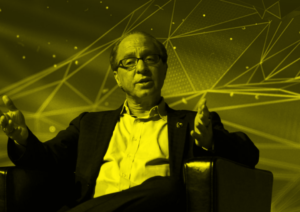Futurist and AI expert Ray Kurzweil speaking at a conference, gesturing with both hands. A digital, geometric background reinforces the theme of technological advancement and the singularity.